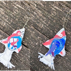 May include: Two handmade fabric fish ornaments with red, blue, and purple paint details. The fish have white fabric bodies and frayed edges. Each fish has a button eye.