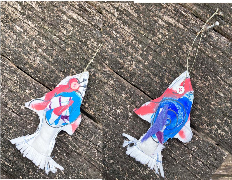 May include: Two handmade fabric fish ornaments with red, blue, and purple paint details. The fish have white fabric bodies and frayed edges. Each fish has a button eye.