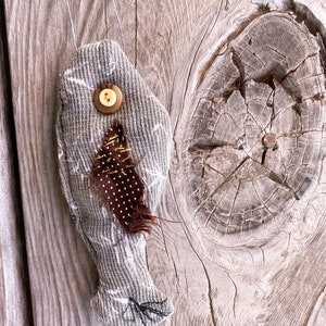 May include: A gray fabric fish ornament with a brown feather and a button for an eye. The ornament is hanging from a string on a wooden background.