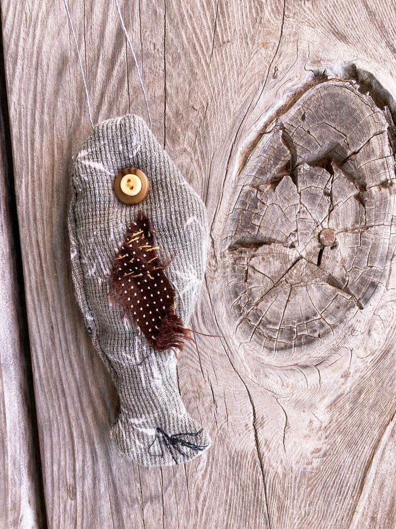 May include: A gray fabric fish ornament with a brown feather and a button for an eye. The ornament is hanging from a string on a wooden background.