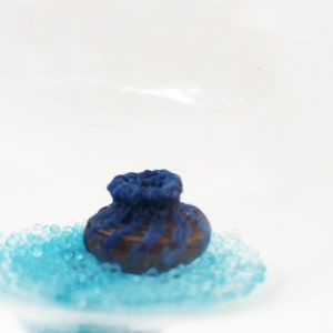 May include: A small blue and brown ceramic pot sits on a bed of light blue iridescent beads. The pot has a rounded shape and a slightly flared rim.
