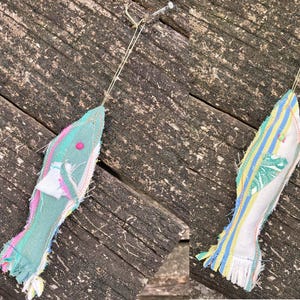May include: Two handmade fabric fish ornaments hanging from a wooden surface. The fish are made from colorful fabric scraps and have a fringed edge. One fish is green and pink, the other is white, yellow, and blue.