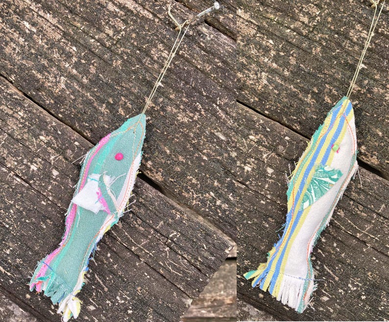 May include: Two handmade fabric fish ornaments hanging from a wooden surface. The fish are made from colorful fabric scraps and have a fringed edge. One fish is green and pink, the other is white, yellow, and blue.