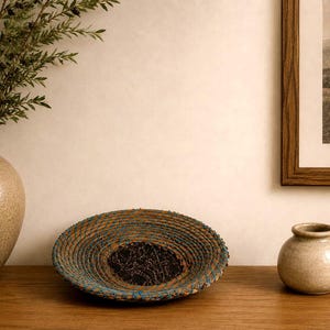 May include: A shallow, round, coiled basket bowl featuring a dark brown center and natural tan fibers wrapped with vibrant blue thread. The bowl is positioned between a tall, speckled beige vase and a small, round, speckled beige pot.