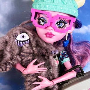 May include: A doll with pink skin, purple hair, and pixelated pink glasses. She wears a teal knit hat with yellow horns and a gray furry shawl with an eye design. The doll has blue earrings and is holding a gray boombox.
