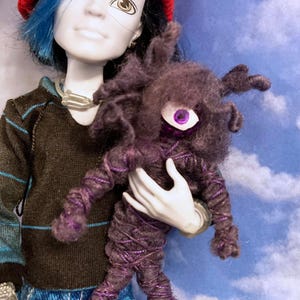May include: A doll with blue hair wearing a red beanie and a brown sweater holds a purple and black yarn creature with a single purple eye.