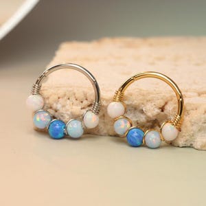 Op de afbeelding: Twee zilveren en gouden oorringen met witte en blauwe opaal kralen. De oorringen liggen op een lichtbruin oppervlak.