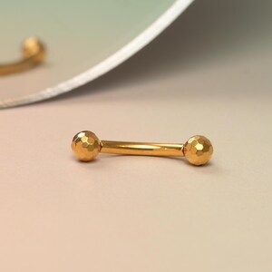 Könnte beinhalten: Goldfarbener Barbell mit strukturierten Kugelenden. Der Barbell ist gebogen und hat eine glänzende Oberfläche.