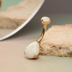 Op de afbeelding: Gouden navelring met twee witte opaalstenen. De grotere steen is druppelvormig.