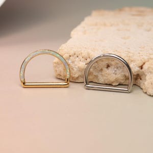 Puede incluir: Dos piezas de joyería corporal en forma de D. Una es dorada con una incrustación de ópalo blanco, la otra es plateada. Ambas se exhiben sobre un fondo neutro con una piedra texturizada.