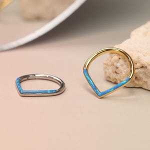 Op de afbeelding: Twee decoratieve ringen. Eén is zilverkleurig met een blauwe opaal inleg in een V-vorm. De andere is goudkleurig met een blauwe opaal inleg in een V-vorm. De ringen worden op een neutrale achtergrond gepresenteerd.