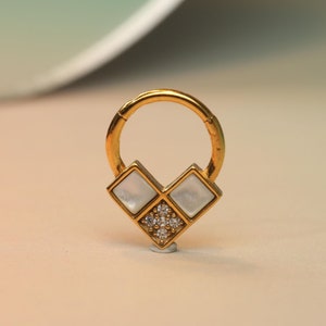 Puede incluir: Pendiente de aro dorado con un dije en forma de corazón blanco y dorado. El dije tiene un diseño geométrico con pequeños diamantes de imitación transparentes.