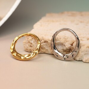 May include: Two textured metal rings, one gold and one silver, are displayed on a light-colored surface. The rings have a hammered or dimpled appearance, adding a unique texture to their design. The background is a soft gradient of green and beige.