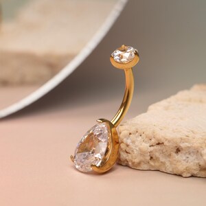 May include: A gold-colored belly button ring with a pear-shaped, clear gemstone and a smaller, round gemstone. The curved bar is gold, and the gemstones are set in gold bezels. The jewelry is displayed on a textured, beige surface.
