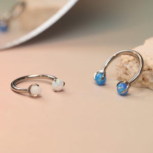 Puede incluir: Dos piercings plateados en forma de herradura con orbes de ópalo en cada extremo. Un anillo tiene orbes blancos y el otro azules. Los anillos se muestran sobre una superficie rosa claro con un fondo neutro.