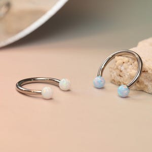 Puede incluir: Dos piezas de joyería corporal plateadas en forma de herradura, cada una con cuentas opalinas blancas y azul claro. Las joyas se exhiben sobre una superficie de color beige claro con un fondo neutro. Probablemente para piercings en orejas, nariz o labios.