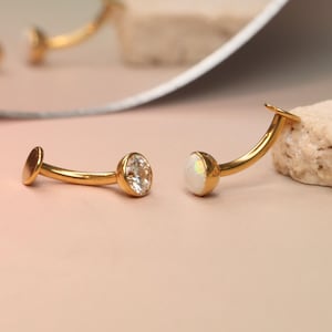 Puede incluir: Dos pendientes dorados curvados, cada uno con un disco plano y una gema. Uno tiene una piedra clara y facetada, el otro una piedra blanca opalescente. Se presentan sobre una superficie rosa claro, con un espejo en el fondo.