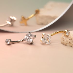 Puede incluir: Piercings para el ombligo con piedras de circonita cúbica transparente. Un anillo es plateado con una piedra cuadrada, y el otro es dorado con una piedra en forma de estrella. Las joyas se exhiben sobre un fondo rosa y beige.