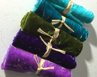 May include: Five rolls of velvet fabric in various colors, including teal, green, purple, and a darker shade of purple. The fabric is tied with twine and arranged in a stack.