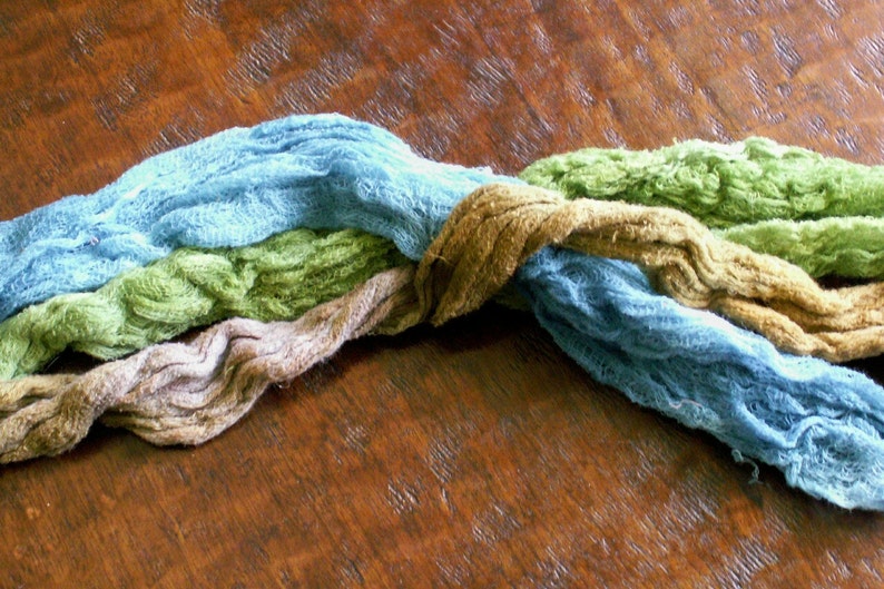 K&ouml;nnte beinhalten: Vier Str&auml;nge Stoff in den Farben Blau, Gr&uuml;n, Braun und Beige sind miteinander verwoben. Der Stoff ist weich und texturiert, und die Farben sind ged&auml;mpft und erdig.