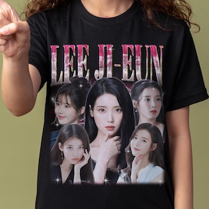 May include: Black t-shirt featuring a vintage-style graphic of Lee Ji-eun, with the text "LEE JI-EUN" in a pink and white gradient. The graphic includes multiple images of the singer. The person is pointing to the left.