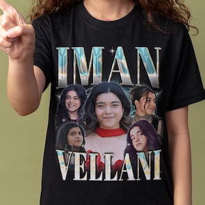 May include: Black t-shirt featuring the name "IMAN VELLANI" in a stylized font, with a collage of images of a young woman. The shirt is worn by a person with long, curly brown hair.