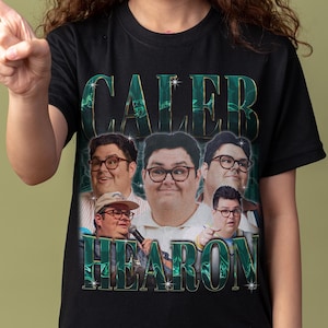 以下が含まれることがあります： 黒いTシャツ。画像と「Caleb Hearon」の文字が緑色の葉っぱのようなフォントでコラージュされています。長いウェーブのかかった茶色の髪の人が、そのTシャツを指さしています。