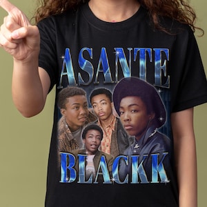 May include: Black t-shirt featuring the text "ASANTE BLACKK" in a blue gradient font. The shirt displays a vintage-style image of a group of young men. The background is a solid olive green.