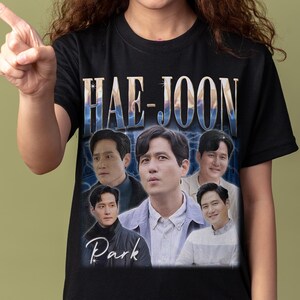 Könnte beinhalten: Schwarzes T-Shirt mit einer Grafik von mehreren Männern und dem Namen "HAE-JOON" in einer stilisierten Schriftart. Das Wort "Park" ist unter den Bildern aufgedruckt. Die Person im Bild zeigt nach links.
