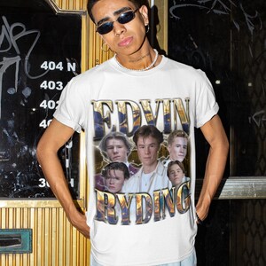 Op de afbeelding: Wit T-shirt met een goud-blauwe afbeelding van de tekst "EDVIN RYDING" en een collage van zes jonge mannen. Het T-shirt wordt gedragen door een persoon met een zonnebril en een lichtblauwe jeans.