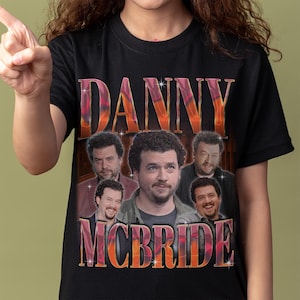 May include: A black short-sleeved t-shirt featuring a graphic print of the actor Danny McBride. The design includes the name DANNY MCBRIDE in large, stylised, orange and pink gradient lettering, surrounding five distinct portraits of the actor.