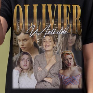 May include: A black t-shirt featuring a large gold-toned graphic print with the text OLLIVIER and Mathilde in script. The design includes a collage of four photographic portraits of a person in various outfits and poses against a city background.