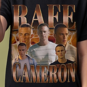 May include: A black t-shirt featuring a graphic print of the name Rafe Cameron in large metallic gold letters at the top and bottom. The center displays four photographic portraits of a young person with short hair against a lightning background.