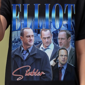 Puede incluir: Una camiseta negra con un estampado gráfico de cuatro hombres. El texto "Elliot Stabler" está impreso en rojo en la parte inferior de la camiseta. Los hombres llevan trajes y corbatas. El fondo es una ciudad.