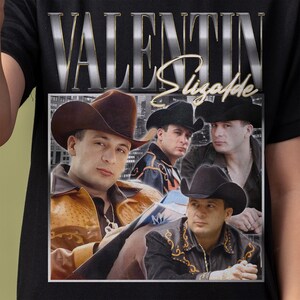 May include: Black t-shirt featuring a graphic of four men in cowboy hats. The image has the text "VALENTIN Elizalde" in silver and gold lettering. The men are in front of a city skyline.