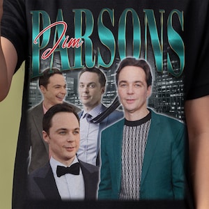Può includere: T-shirt nera con un collage di immagini di Jim Parsons. La parola "PARSONS" è stampata in grandi lettere turchesi, con "Jim" in corsivo. Il design include più immagini dell'attore in vari outfit.