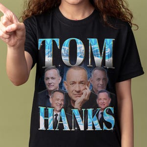 Puede incluir: Una camiseta negra de manga corta con un estampado gráfico del actor Tom Hanks. El diseño incluye el nombre TOM HANKS en letras grandes de color azul metálico y plateado con un patrón de remolinos, rodeando varios retratos del actor.