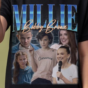 May include: Black t-shirt featuring a graphic of Millie Bobby Brown with the text "MILLIE" in blue and "Bobby Brown" in gold. The design includes multiple images of the actress.