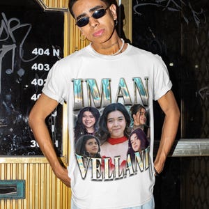 May include: White t-shirt featuring the name "IMAN VELLANI" in a stylized font, with images of the actress. The shirt has a vintage design. The person is wearing sunglasses and a pearl necklace.