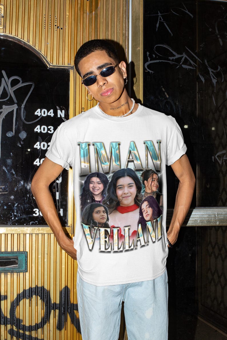 May include: White t-shirt featuring the name "IMAN VELLANI" in a stylized font, with images of the actress. The shirt has a vintage design. The person is wearing sunglasses and a pearl necklace.