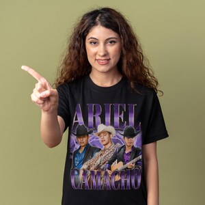May include: Black t-shirt featuring a vintage-style graphic of three men in cowboy hats, with the name "ARIEL CAMACHO" in purple text. The design includes a purple lightning bolt effect behind the figures.