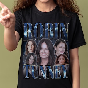 May include: A black short-sleeve t-shirt featuring a graphic print of the actor Robin Tunney. The design includes the name ROBIN TUNNEY in large blue metallic-style lettering with star accents, surrounding five different photographic portraits of the actor.