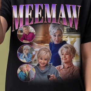 May include: A black t-shirt with the word "MEEMAW" printed in purple and white letters. The shirt features a collage of images of five people, each in a circular frame. The people are all smiling and appear to be related.