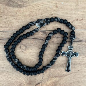 May include: A black rosary with round beads and a silver crucifix. The beads are linked by a black and white cord. The crucifix shows a detailed depiction of Jesus Christ and the letters INRI.