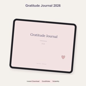 Peut inclure: Une tablette numérique affichant la page de couverture d'un journal de gratitude 2026 avec un fond rose pâle, du texte sombre et une petite icône de cœur souriant. L'écran indique le titre Gratitude Journal, l'année 2026 et des applications.