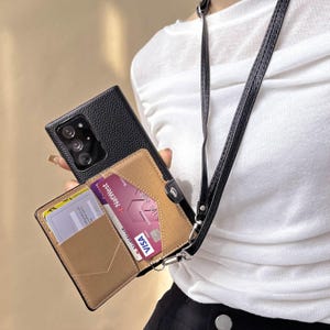 Könnte beinhalten: Schwarze Leder-Handyhülle mit einem beigefarbenen Kartenhalter, der Kreditkarten enthält. Die Hülle hat einen schwarzen Riemen zum Umhängen. Die Handyhülle ist geöffnet und zeigt die Karten im Inneren.