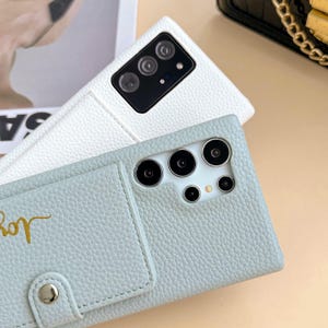 May include: Two leather phone cases, one white and one light blue, are displayed. The light blue case has a card slot and gold script that reads "Joy". Both cases have multiple camera lenses.