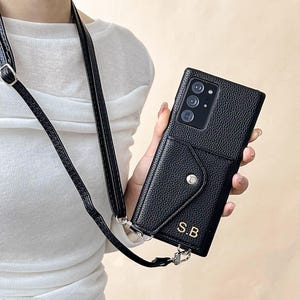 Könnte beinhalten: Schwarze Leder-Handyhülle mit Umhängeband. Die Hülle hat eine strukturierte Oberfläche, einen Klappenverschluss mit Druckknopf und eine kleine Tasche. Die Initialen "S.B" sind goldfarben auf der Vorderseite aufgedruckt. Die Hülle ist für ein Smartphone.