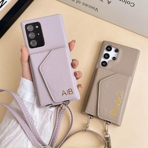 May include: Two leather phone cases with cardholder pockets and detachable straps. One case is lavender with the initials "AIB" and the other is taupe with the name "Phoebe" embroidered in gold.