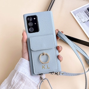 May include: A pale blue leather phone case with a strap. The case features a card slot, a gold ring holder, and the initials "K.L." embossed on the front. Designed to fit a smartphone.